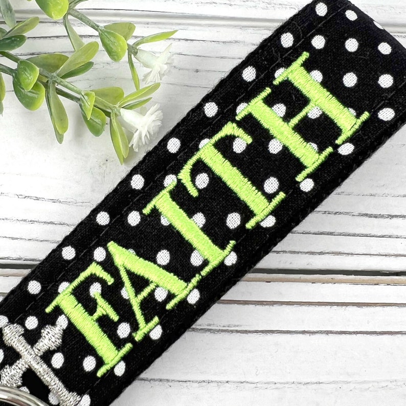 Pu&ograve; includere: Portachiavi in tessuto nero a pois bianchi con la parola "FAITH" ricamata in verde.
