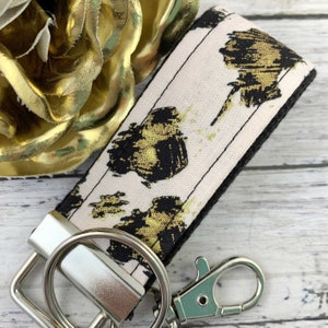May include: A pink and black key chain with a gold abstract print. The key chain has a silver key ring and a silver clip.