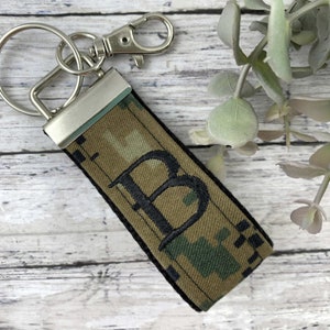 May include: A camouflage patterned key chain with a black embroidered letter "B" on it. The key chain has a silver metal ring and a silver metal clasp.