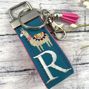 May include: Teal blue fabric keychain with a pink trim. The keychain features an embroidered llama with a colorful saddle and the letter "R" in white. The keychain has a silver metal ring and a silver metal clasp.