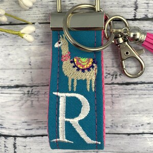 May include: Teal blue fabric keychain with a white embroidered llama and the letter "R" in white. The keychain has a silver metal ring and a silver metal clasp.