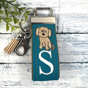 May include: Teal blue fabric keychain with a brown dog embroidered on it. The keychain has a silver metal ring and a brown tassel. The letter "S" is embroidered in white on the fabric.