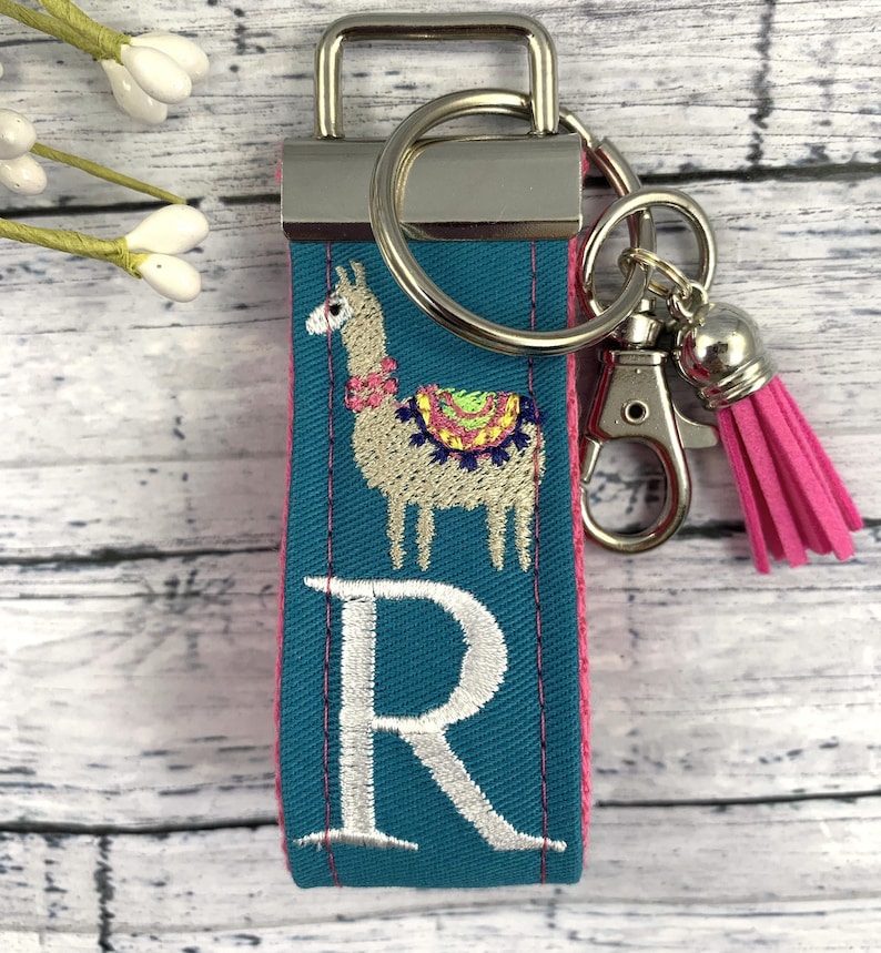 May include: Teal blue fabric keychain with a white embroidered letter R and a llama with a colorful saddle. The keychain has a silver key ring and a pink tassel.