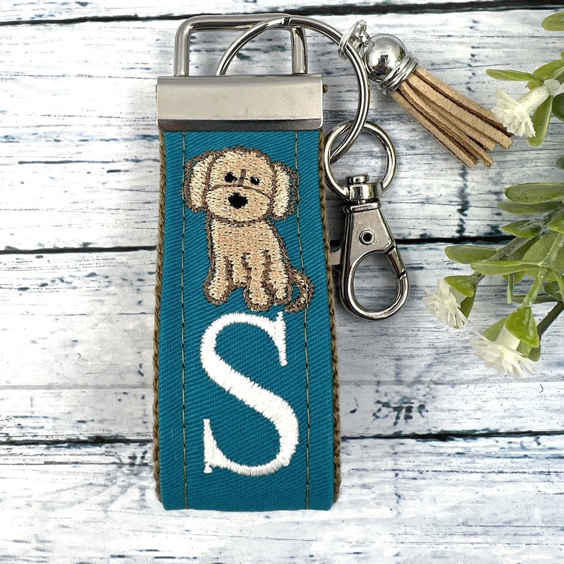 May include: Teal blue key chain with a brown dog and the letter "S" embroidered on it. The key chain has a silver metal clasp and a brown tassel.