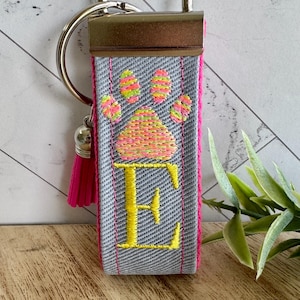 May include: A gray fabric keychain with a pink border, a yellow embroidered letter "E", and a colorful paw print design. The keychain has a silver metal clasp, a pink tassel, and a silver charm.