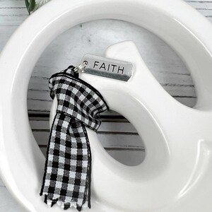 Pu&ograve; includere: Un segno di pace in ceramica bianca con un nastro a quadri bianco e nero attaccato. Il nastro ha un ciondolo d'argento con la parola "FAITH" incisa.