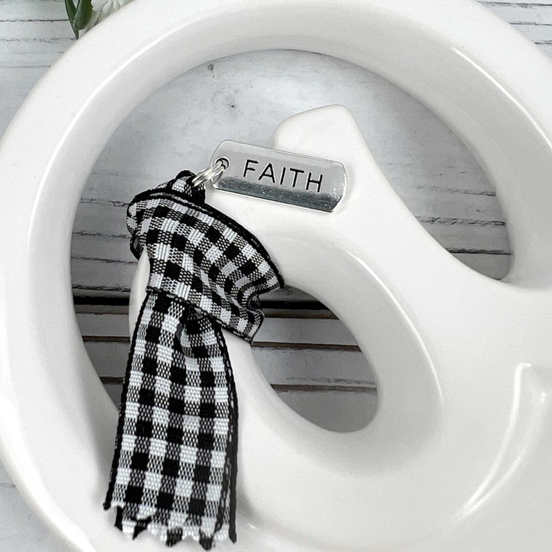 Pu&ograve; includere: Un segno di pace in ceramica bianca con un nastro a quadri bianco e nero attaccato. Il nastro ha un ciondolo d'argento con la parola "FAITH" incisa.