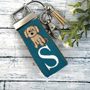 May include: Teal blue key chain with a small embroidered dog and the letter "S" in white. The key chain has a silver metal clasp and a brown tassel.