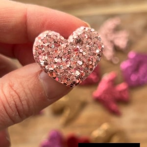 Peut inclure: Un embellissement en forme de cœur rose pailleté. Le cœur est fait d'un matériau scintillant et a une couleur rose clair. Le texte "Coeur rose poudré" est visible sur l'image.