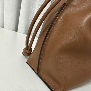 May include: Close-up view of a brown leather drawstring bucket bag. The bag features a simple, minimalist design with clean lines and a subtle, dark brown stitching detail along the edges. The drawstring closure is made of the same brown leather as the bag itself, adding to its cohesive aesthetic. The leather appears to be of high quality, with a smooth, slightly textured surface that suggests durability and sophistication. The bag's shape is slightly slouchy, giving it a relaxed and casual feel, while still maintaining a polished look. This versatile bag would be suitable for everyday use or more formal occasions.