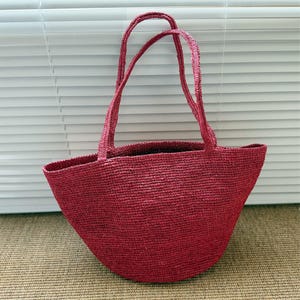 May include: A red woven tote bag with two handles. The bag has a rounded bottom and a wide opening. The material appears to be a natural fiber, and the bag is a solid, vibrant red color. The handles are the same color as the bag.