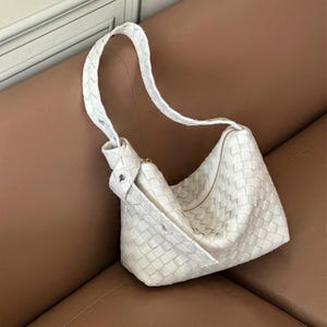 Può includere: Una borsa a tracolla bianca in pelle intrecciata con una forma curva e una singola tracolla. La borsa presenta una chiusura con cerniera e un dettaglio annodato alla base della tracolla. La borsa è su una superficie in pelle marrone.