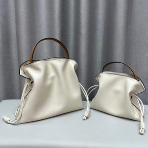 May include: Two off-white leather handbags with knotted drawstrings and tan leather handles. The larger bag is positioned to the left, and the smaller bag is to the right. Both bags have a soft, gathered design.