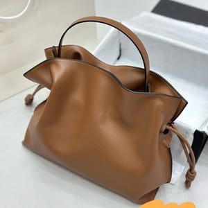 May include: A brown leather handbag with a top handle and drawstring closure. The bag is soft and slouchy, with a relaxed, casual look.  The leather appears smooth and supple. The bag's simple design is highlighted by its rich brown color and subtle stitching. This stylish tote is perfect for everyday use.