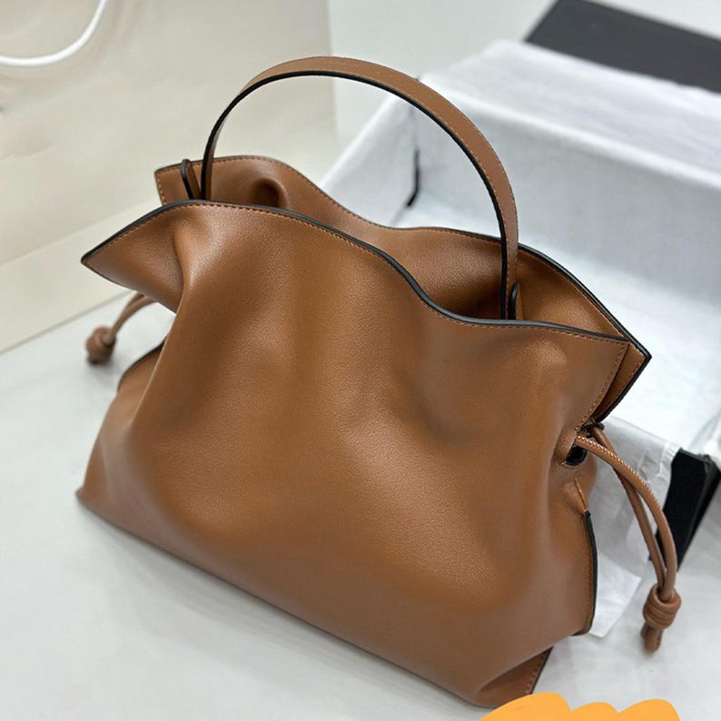 May include: A brown leather handbag with a top handle and drawstring closure. The bag is soft and slouchy, with a relaxed, casual look.  The leather appears smooth and supple. The bag's simple design is highlighted by its rich brown color and subtle stitching. This stylish tote is perfect for everyday use.