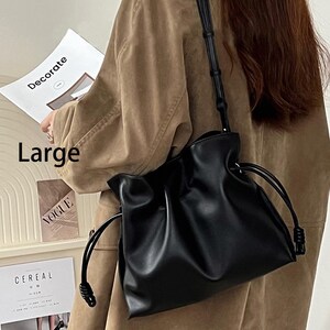 May include: A black, large, drawstring bucket bag made of faux leather. The bag has a relaxed, slouchy silhouette and features knotted drawstring closures.  The bag is shown hanging from a shoulder strap.