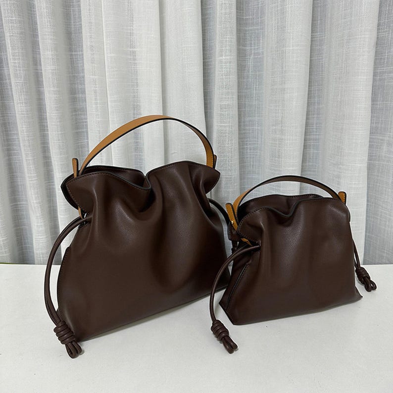 May include: Two brown leather bucket bags with tan leather handles and drawstring closures. The larger bag is positioned behind and slightly to the left of the smaller bag. Both bags have a gathered, slouchy appearance.  The bags are displayed against a neutral background.