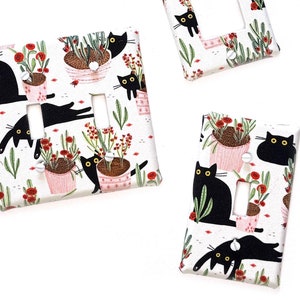 May include: Three light switch covers with a black cat and plant design. The covers are white with a pink, red, and green floral pattern. The cats are black with yellow eyes. The switch covers are a decorative way to add a touch of whimsy to any room.