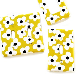 Puede incluir: Tres tapas de interruptor de luz amarillas con un patrón floral blanco. Las tapas tienen un interruptor simple y un interruptor doble.