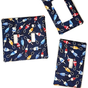 May include: Three rectangular wall switch plates featuring a dark navy blue background with a repeating pattern of colorful nautical buoys in shades of red, white, yellow, and light blue. The set includes one double toggle plate and two single plates.