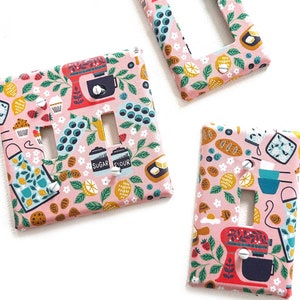 以下が含まれることがあります： キッチンをテーマにしたパターンが施されたピンクのライトスイッチカバーが3つ。パターンには、スタンドミキサー、レモン、ブルーベリー、そして「砂糖」と「小麦粉」の文字が描かれています。