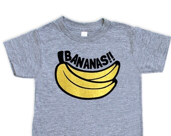 Op de afbeelding: Een grijs T-shirt met korte mouwen met een zwarte en gouden afbeelding van bananen en de tekst "BANANAS!" gedrukt op de voorkant.