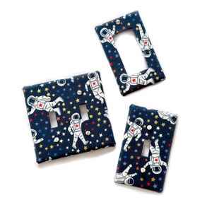 May include: Three astronaut-themed light switch covers. The covers are navy blue with white stars and yellow accents. The astronauts are white with red accents. The covers are for standard light switches.