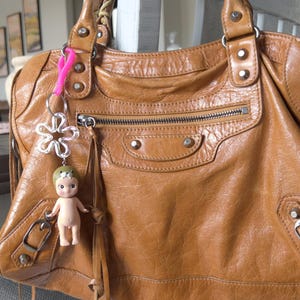 May include: A brown leather handbag with a zippered pocket and a silver metal flower charm. The bag has a long strap and a small doll keychain.