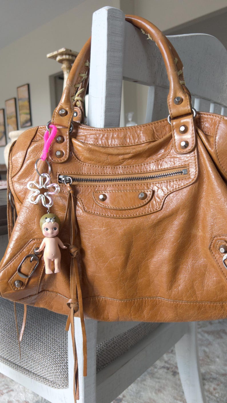 May include: A brown leather handbag with a zippered pocket and a silver metal flower charm. The bag has a long strap and a small doll keychain.