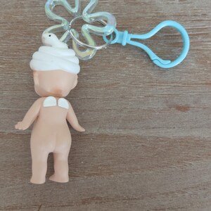 May include: A small, nude-colored doll with a white towel on its head. The doll is attached to a blue plastic keychain with a clear plastic flower charm.