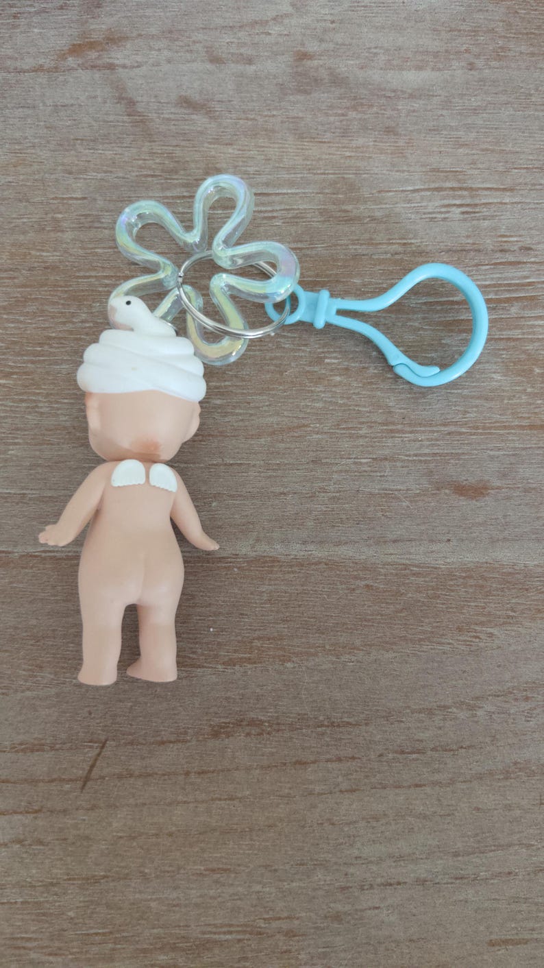 May include: A small, nude-colored doll with a white towel on its head. The doll is attached to a blue plastic keychain with a clear plastic flower charm.