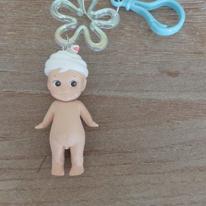 May include: A small, nude, plastic doll with a white towel on its head. The doll is attached to a silver key ring with a blue plastic clip.