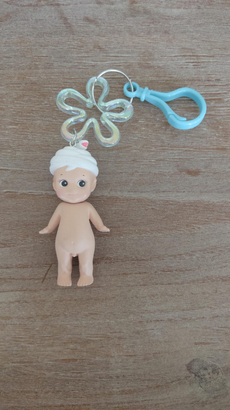 May include: A small, nude, plastic doll with a white towel on its head. The doll is attached to a silver key ring with a blue plastic clip.
