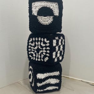 May include: Three stacked, handmade crochet blocks in black and white. Each block features a different geometric design, including a circle, floral pattern, and striped pattern. The blocks are likely intended for baby toys or nursery decor.
