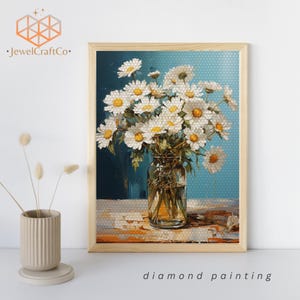 May include: A framed diamond painting featuring a bouquet of white daisies in a clear glass vase. The daisies are arranged in a loose, natural style, and the vase is sitting on a textured surface. The background is a light blue color.