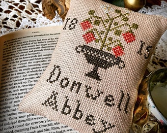 May include: A cross-stitch pattern for a cushion featuring a black bird with a red berry bush and the text "Donwell Abbey". The pattern is inspired by Jane Austen's novels.