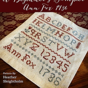 May include: A beginner's sampler cross stitch pattern featuring the alphabet, numbers, and the text "Ann Fox 1736". The pattern is stitched in red and blue thread on white Aida cloth.