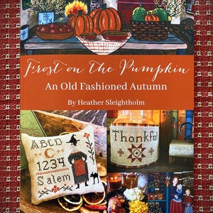 Puede incluir: Una portada de libro con temática otoñal. La portada presenta una pintura de calabazas, manzanas y una chimenea. El título del libro es "Frost on the Pumpkin: An Old Fashioned Autumn" de Heather Sleightholm. La portada también incluye un collage de patrones de punto de cruz, incluyendo una calabaza, una bruja y un letrero de agradecimiento.