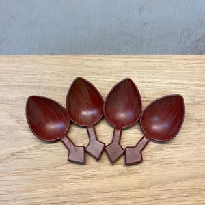 Puede incluir: Cuatro cucharas de color granate dispuestas en fila. Cada cuchara tiene un cuenco en forma de lágrima y un mango rectangular. Las cucharas son de un material liso y están sobre una superficie de madera clara.