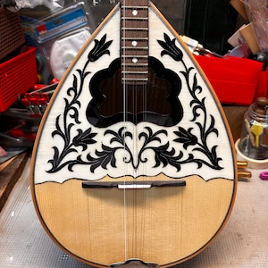 May include: A pear-shaped stringed instrument with a light wood body and a decorative white and black inlaid top. The instrument features a dark fretboard and strings, with a metal tailpiece. The instrument is likely a mandolin or similar folk instrument.