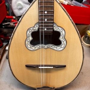 May include: A close-up of a mandolin with a light wood grain body and a dark brown fretboard. The instrument features a decorative, cloud-shaped inlay around the sound hole and a dark brown bridge. The strings are visible, stretched across the body.