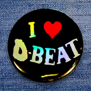 Op de afbeelding: Een zwarte knop met een holografisch regenboog-effect. De knop zegt "Ik hou van D-Beat" met een rood hart in het midden.