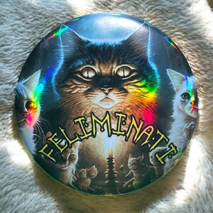 Peut inclure: Un bouton holographique avec un chat noir aux yeux jaunes et le texte "FELIMINATE" en lettres dorées. Le chat est entouré de chats plus petits et d'une bougie.