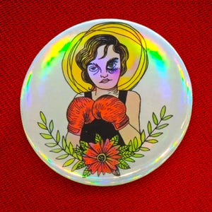 Peut inclure: Un bouton rond et holographique avec une illustration stylisée d'une personne portant des gants de boxe orange et un haut noir. La personne est encadrée d'un halo jaune et d'un motif floral. La personne a un œil au beurre noir.