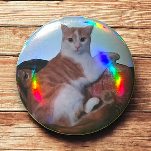 Peut inclure: Un gros plan sur un bouton rond et iridescent avec une photographie d'un chat blanc et orange assis sur une surface à motifs rouge et blanc.