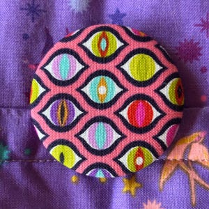 Op de afbeelding: Een roze stoffen knoop met een herhalend patroon van kleurrijke ogen. De ogen zijn zwart omrand en hebben verschillende gekleurde pupillen en iris.