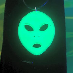 Könnte beinhalten: Ein leuchtend grüner Alien-Anhänger an einer schwarzen Kordel. Der Anhänger hat die Form eines Alien-Kopfes mit großen ovalen Augen und einem kleinen Mund. Der Anhänger leuchtet hell vor einem dunklen Hintergrund.