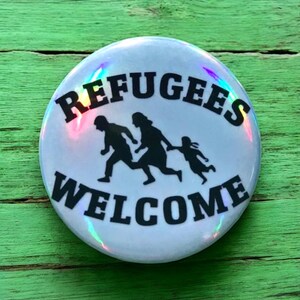 May include: A white button with a rainbow iridescent finish. The button features black text that reads "Refugees Welcome" and a silhouette of a family running.