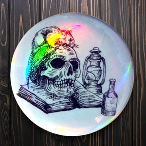 Puede incluir: Un plato de cerámica blanco con una ilustración en blanco y negro de una calavera con una rata sentada encima. La ilustración también incluye un libro, una linterna y una botella.