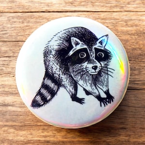 Peut inclure: Un bouton blanc avec une illustration en noir et blanc d'un raton laveur. Le bouton a une finition irisée.